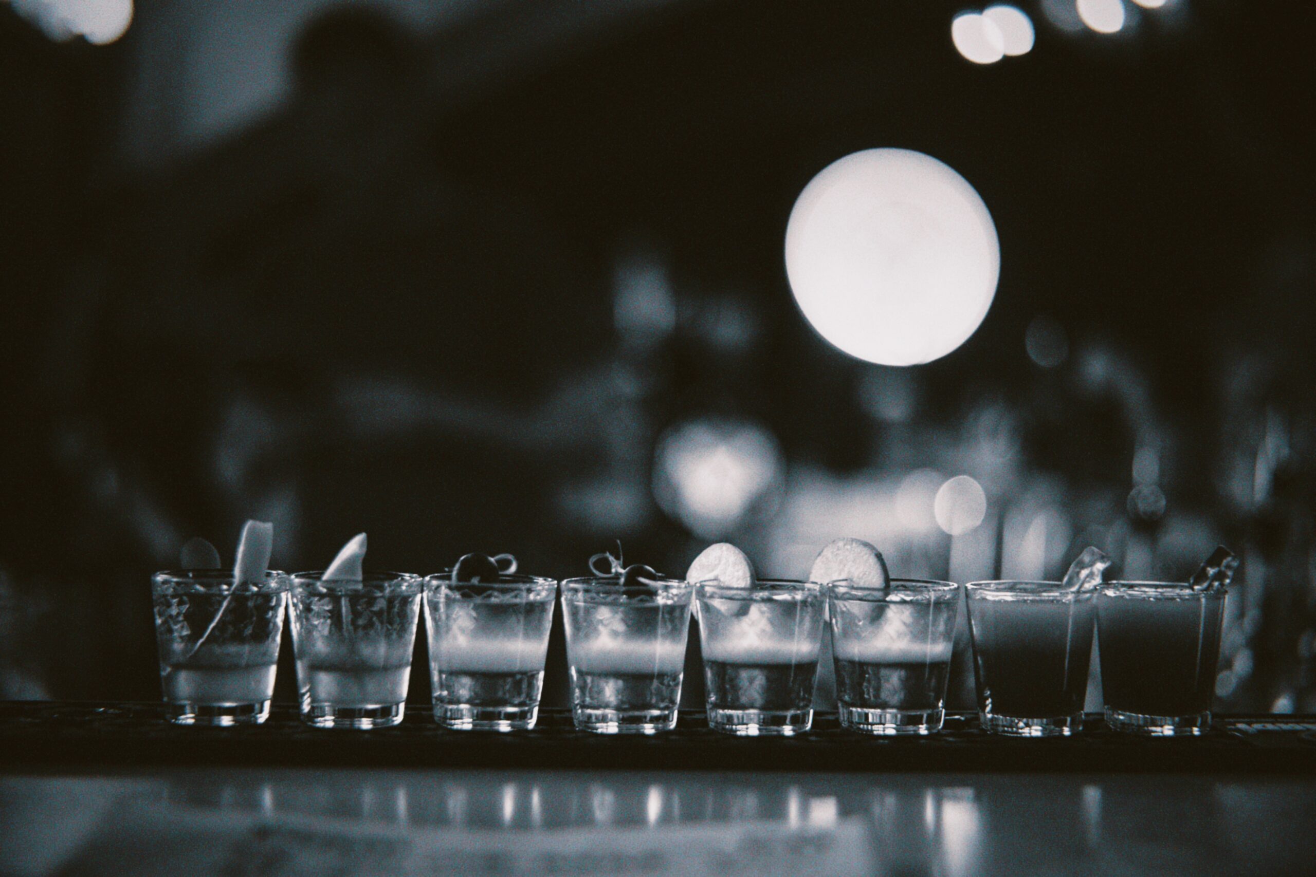 Série de verres à shot remplis et alignés sur un comptoir de bar. Chaque verre est garni d'une tranche de citron vert. L'arrière-plan est flou avec des lumières tamisées, créant une ambiance de bar.